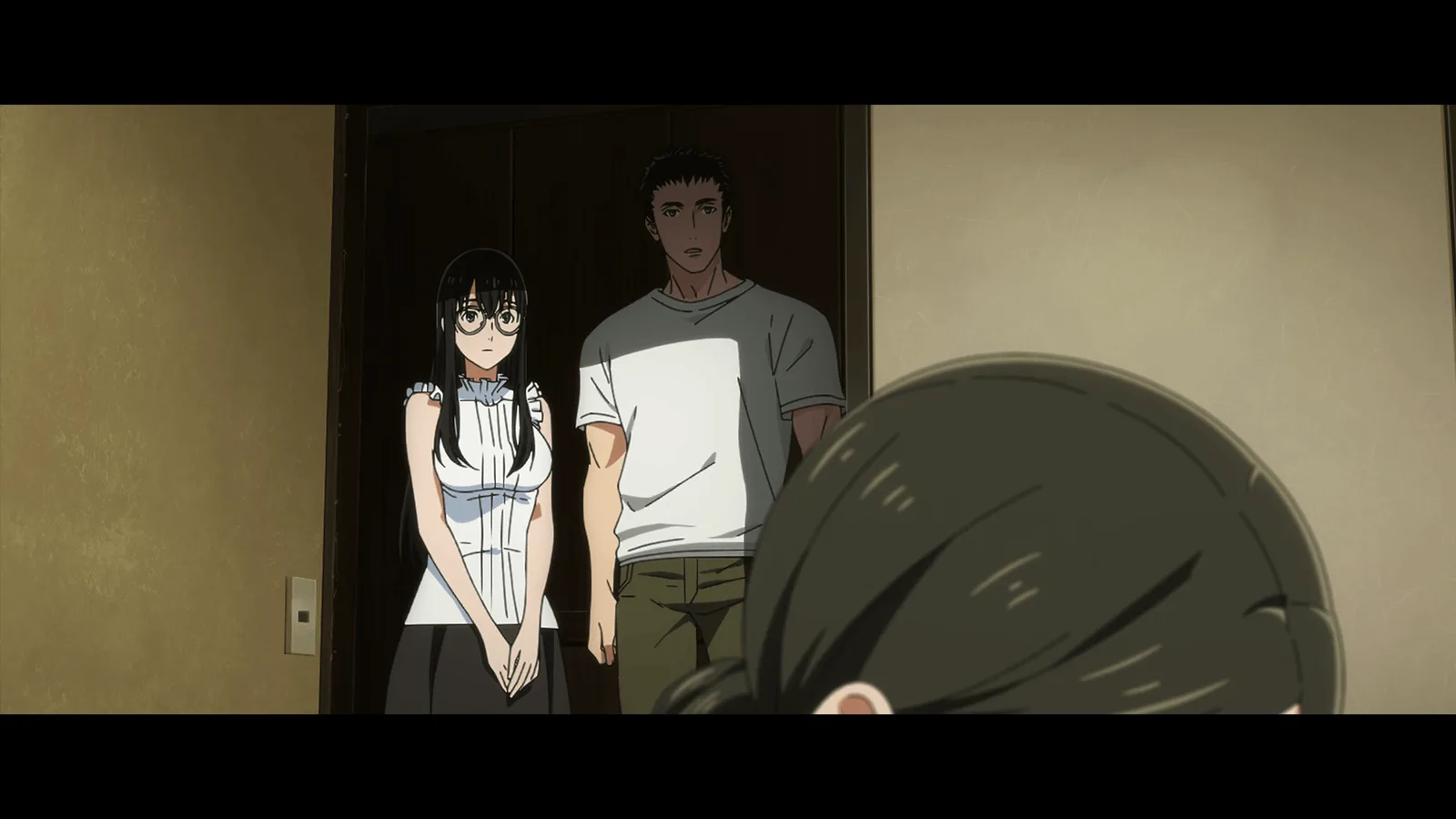 Une jeune femme brune à lunettes et un grand homme en t-shirt se tiennent devant une porte sombre, observés par un personnage vu de dos au premier plan.